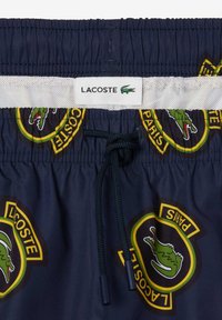 Bañador azul marino con cintura ajustable por cordón, presenta los logos de Lacoste en amarillo y verde, y un forro de malla para mayor comodidad.