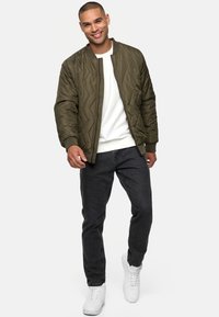 Chaqueta bomber acolchada en verde oliva con puños de canalé, llevada sobre un sudadera blanca, combinada con jeans oscuros y zapatillas blancas.