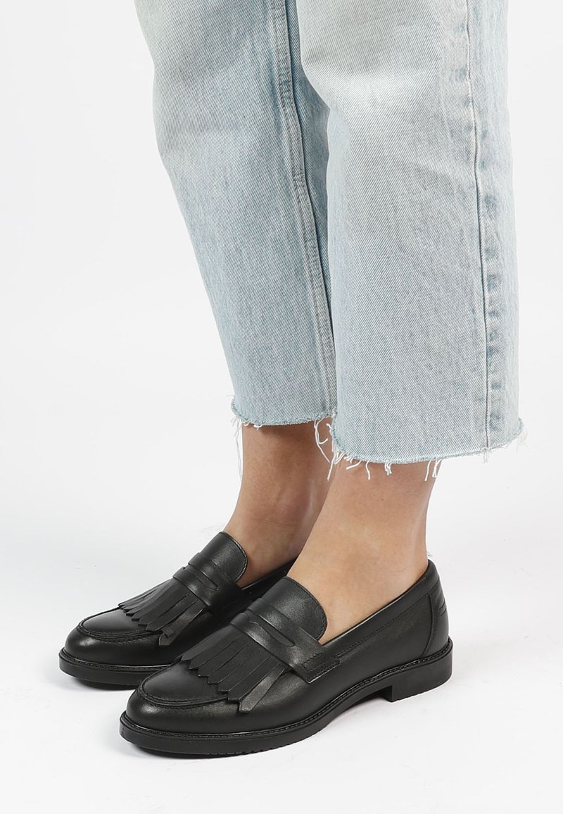 Persoon in zwarte leren loafers met franjes en lichtblauwe cropped jeans met rafelige zoom op een witte achtergrond.