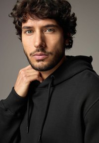 Schwarzer Kapuzenpullover aus weichem Stoff, mit verstellbaren Kordeln und gerippten Bündchen. Nahaufnahme eines männlichen Modells mit lockigem Haar.