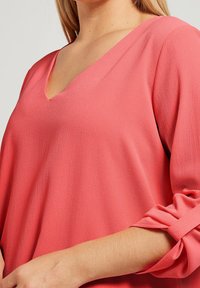 Blusa corallo a V con maniche tre quarti, caratterizzata da un tessuto texturizzato e dettagli annodati alle maniche. Design leggero e dalla vestibilità ampia.