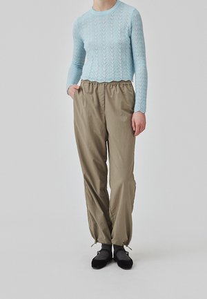 Personne portant un haut texturé bleu clair à manches longues, un pantalon beige ample avec des poignets élastiques, des chaussettes grises et des chaussures plates noires, debout avec une main dans la poche.