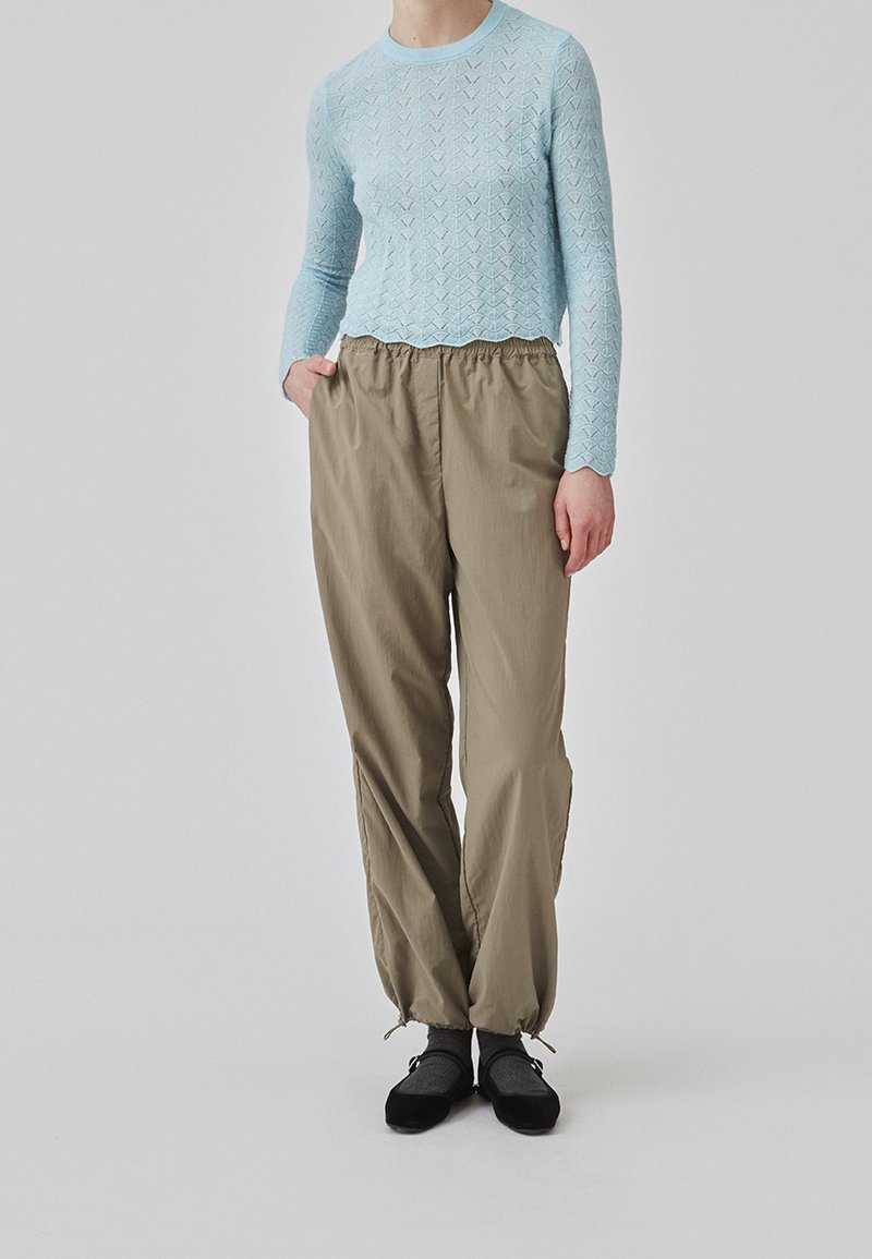Personne portant un haut texturé bleu clair à manches longues, un pantalon beige ample avec des poignets élastiques, des chaussettes grises et des chaussures plates noires, debout avec une main dans la poche.