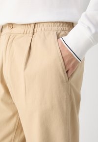Osoba ubrana w beżowe spodnie z elastycznym pasem i guzikiem, ręka w kieszeni, biała koszula z długim rękawem i paskowanym mankietem.