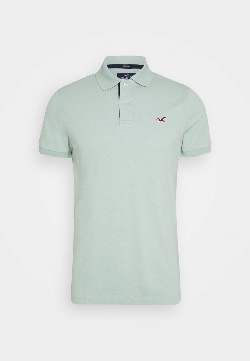 Polo à manches courtes vert clair avec col, deux boutons blancs, et petit logo d'oiseau rouge sur le côté gauche de la poitrine.