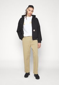 Black hooded jacket with a zip, beige trousers, and a white t-shirt featuring gold text. Footwear is black sneakers. Plain background.