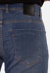 Vaqueros de mezclilla azul oscuro con un bolsillo trasero con costuras naranjas y una etiqueta de cuero encima. La tela tiene un acabado texturizado.