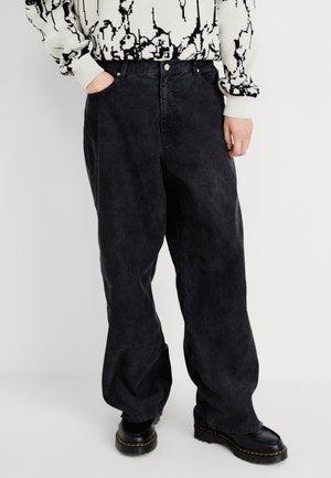 Person trägt übergroße schwarze gewaschene Jeans, schwarze Plateauschuhe und einen weißen Pullover mit einem schwarzen abstrakten Muster.
