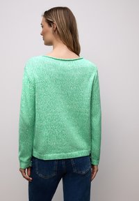 Femme vue de dos portant un pull en maille vert à manches longues et un jean bleu sur un fond clair uni.