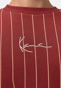Camiseta de algodón a rayas rojas con cuello redondo y firma bordada en blanco en el pecho, destacando un diseño moderno y elegante.
