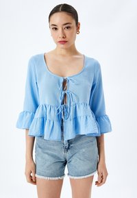 Lichtblauwe blouse met een ronde halslijn, een strikdetail aan de voorkant en gerafelde mouwen. Gecombineerd met denim shorts met een rafelige zoom.
