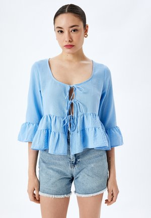 Blusa azzurra chiara con scollatura rotonda, dettaglio di nodo frontale e maniche volanti. Abbinata a pantaloni corti in denim con orlo sfrangiato.