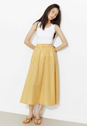 Femme appuyée contre un mur blanc, portant un top blanc sans manches, une jupe midi vichy jaune et des sandales marron à talons.