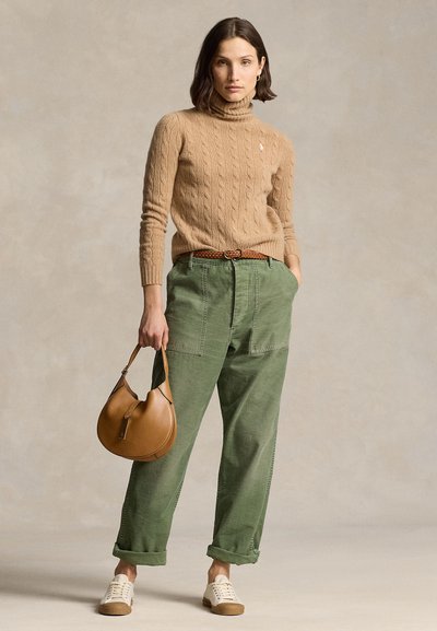 Femme portant un pull à col roulé beige en maille torsadée, pantalon vert avec revers, baskets blanches, tenant un sac à main en cuir marron, debout devant un fond neutre.