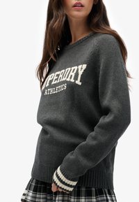 Szary sweter dzianinowy z okrągłym dekoltem, z napisem "Superdry Athletics" w białym kolorze. Mankiety z prążkowanej dzianiny mają biało-czarne paski.