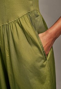 Chiusura verde a costine con vita arricciata e tasche laterali; texture liscia con leggero lucido, che mette in risalto il dettaglio del tessuto stratificato.