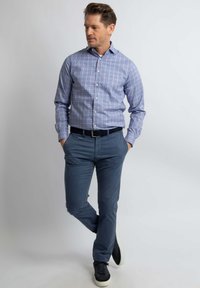 Blauw-witte geruite overhemden met knoopmanchetten, gecombineerd met marineblauwe chinos en zwarte sneakers, op een effen achtergrond.