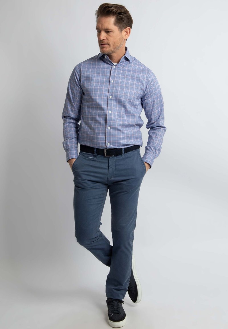 Blauw-witte geruite overhemden met knoopmanchetten, gecombineerd met marineblauwe chinos en zwarte sneakers, op een effen achtergrond.