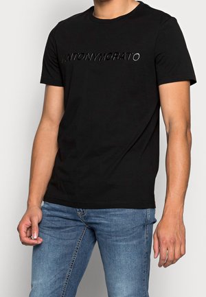 Camiseta negra de algodón con mangas cortas, con el texto en relieve "ANTONY MORATO" en el pecho, combinada con jeans azules.