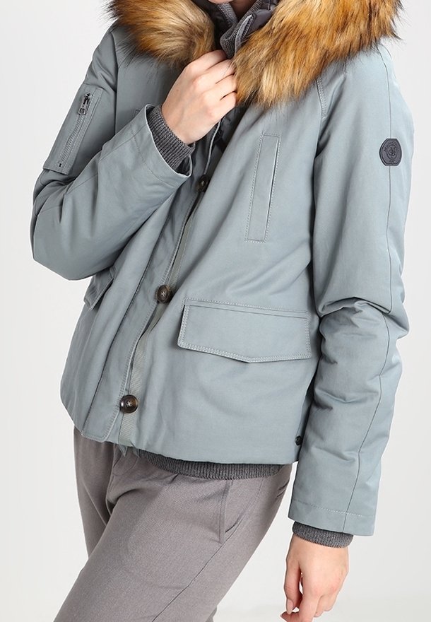Personne portant une veste d'hiver bleu clair avec col en fourrure, fermeture par boutons et fermeture éclair, et poche zippée sur la manche.