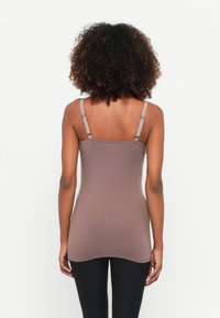 Žena s kovrčavom kosom nosi mauve top na bretele i crne tajice, prikazana sa leđa na bijeloj pozadini.