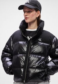 Melna pūkainā jaka ar spīdīgu apdari, augstu apkakli un rāvējslēdzēja aizdari. Iespējo balts logo kreisajā pusē un elastīgas aproces.