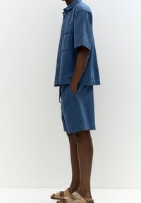 Chemise en coton bleu à manches courtes avec col boutonné et poche poitrine, assortie à un short. Portée avec des sandales beige.