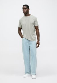 Άνδρας που στέκεται, φορώντας ένα πουκάμισο με μοτίβο και κοντό μανίκι, ανοιχτό μπλε φαρδύ τζιν και λευκά αθλητικά παπούτσια, με το ένα χέρι στην τσέπη.