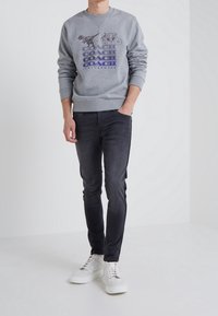 Man i grå sweatshirt med grafik av dinosaurie och vagn, svarta slim fit jeans och vita sneakers, stående med ena handen i fickan.