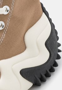 Tan canvas sneaker featuring a textured black rubber sole with jagged edges and a white wavy accent near the midsole. Silver eyelets are visible.