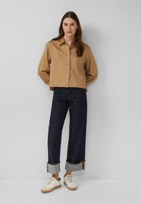 Frau steht vor einer hellgrauen Wand und trägt ein braunes Button-Up-Hemd, dunkle hochgerollte Jeans und weiße Sneakers mit beigen Sohlen.