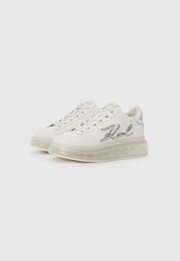 Zapatillas de cuero blancas con suela translúcida, que presentan la palabra "Karl" bordada en plata en el lateral y un diseño de cordones planos.