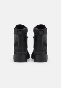 Une paire de bottes noires à la cheville avec des semelles épaisses, en cuir texturé, un col rembourré et des languettes, présentée de dos sur un fond blanc.