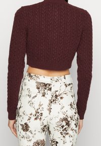 Burgunderfarbener gestrickter kurz geschnittener Pullover mit Zopfmuster und langen Ärmeln, kombiniert mit weißen Hosen mit braunem Blumenmuster.