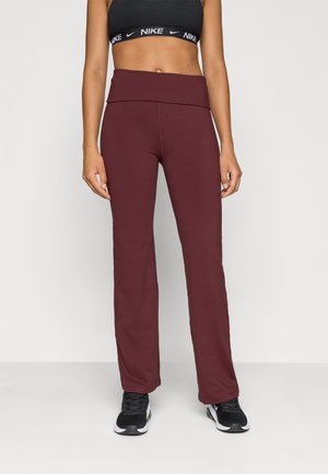 Vrouw draagt een zwarte Nike sport-bh, bordeaux hoge taille wijd uitlopende legging en zwarte Nike sneakers, staand tegen een effen achtergrond.