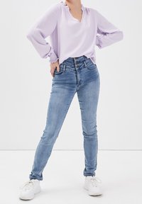 Blusa viola chiaro con scollatura e polsini arricciati, abbinata a jeans skinny azzurri a vita alta e sneakers bianche.