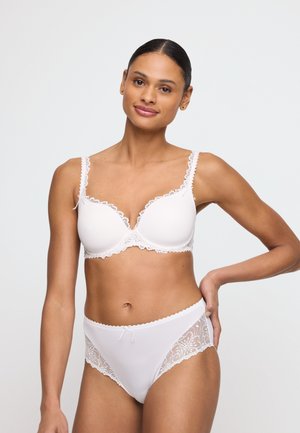 Kobieta w białym biustonoszu z koronkową lamówką i pasujące koronki majtki z wysokim stanem, stojąca z jedną ręką na biodrze na jednolitym tle.