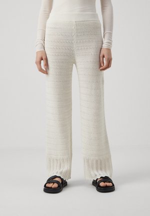 Pantalons en tricot à jambes larges en blanc cassé avec des rayures texturées, dotés d'une taille côtelée et d'un ourlet à franges. Associés à des sandales noires.