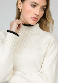 Pull col roulé blanc à côtes avec des bordures noires aux poignets, affichant une coupe décontractée et une texture douce. Le modèle montre le détail des motifs tricotés.
