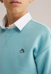 Svetlomodrá mikina s bielym golierom a vyšívaným čiernobielym logom vtáka na ľavej strane hrudníka. Mäkká textúra látky.