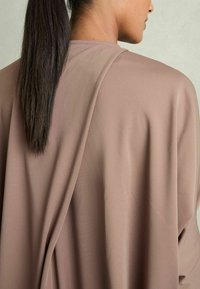 Lös, långärmad topp i taupe-färgad tyg. Slät textur med draperad design, med minimal sömnad längs halsringningen.