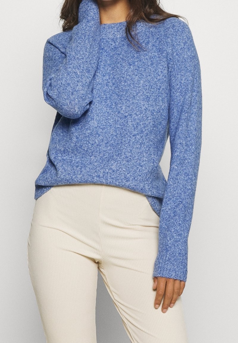 Femme portant un pull bleu texturé à manches longues et un pantalon côtelé beige clair, une main touchant son visage.