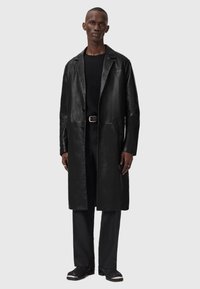 Cappotto di pelle nera con colletto a rever, chiusura con bottoni e maniche lunghe, indossato sopra un maglione nero a maglia e pantaloni neri.