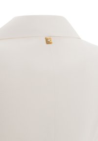 Blusa color crema con colletto classico, dotata di un piccolo logo dorato sul retro e realizzata in un tessuto liscio e texture.