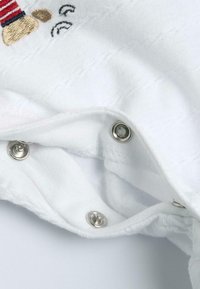 Monosie de tela blanca con cierres de presión plateados, con un diseño bordado que presenta franjas rojas y azules. Patrón de tejido texturizado visible.