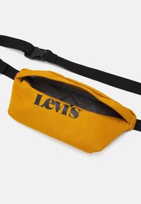 Borsetta Levi's gialla senape con tracolla nera regolabile e cerniera parzialmente aperta su uno sfondo bianco.