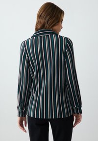 Bluzka z długim rękawem w pionowe paski w kolorze zielonym, czarnym i białym. Posiada kołnierzyk oraz mankiety zapinane na guziki. Gładki materiał i dopasowany krój.