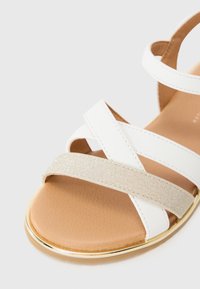 Hvit sandal med kryssreimer, med et glitterende gullbånd. Flat såle med en glatt brun finish og et gullaccent rundt kanten.