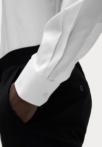Camisa blanca de manga larga con puños abotonados, combinada con pantalones negros. La camisa tiene una textura suave y un corte ajustado.