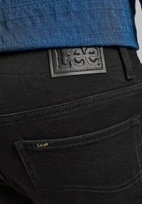 Vaqueros de mezclilla negra con una etiqueta de cuero negro texturizado que presenta la marca "Lee", costuras amarillas en el bolsillo trasero y una camisa azul asomándose por encima.